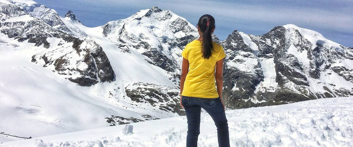 Una persona in piedi su una montagna innevata, con terreno roccioso e cime innevate sullo sfondo.