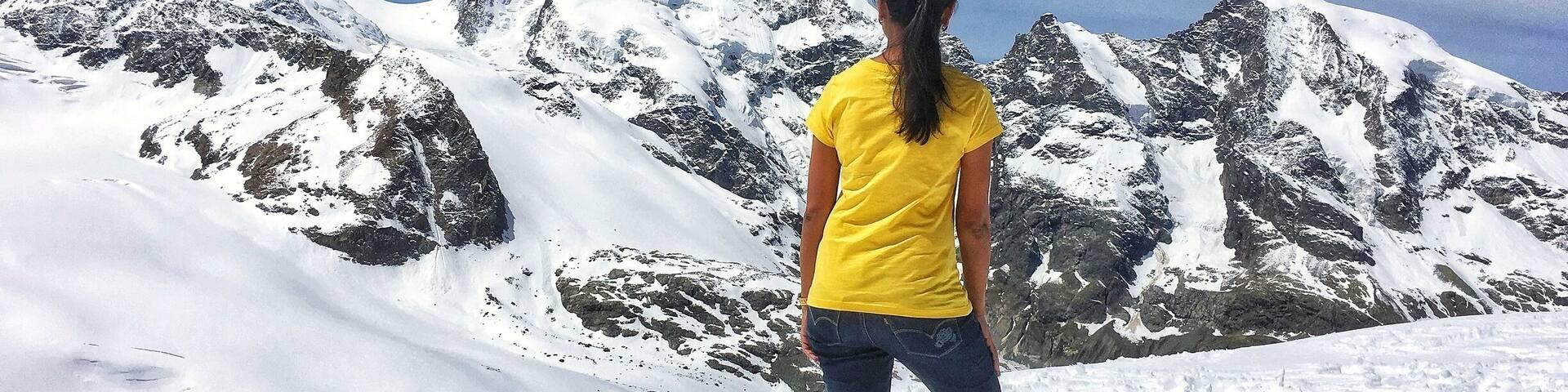 A person standing on a snow-covered mountain with rocky terrain and snow-capped peaks in the background.