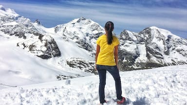 Una persona está de pie sobre una montaña cubierta de nieve, con terreno rocoso y picos nevados al fondo.