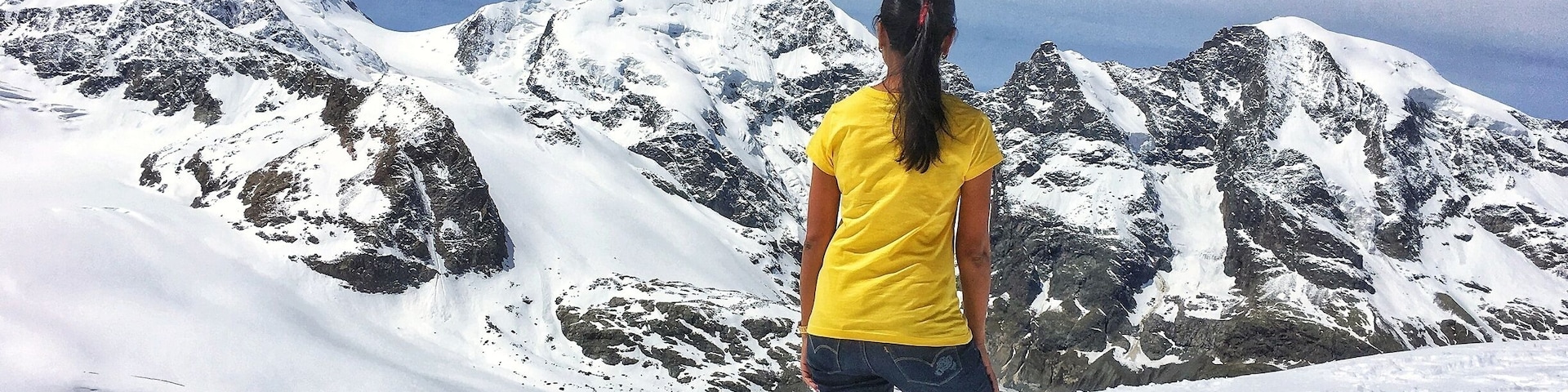Una persona está de pie sobre una montaña cubierta de nieve, con terreno rocoso y picos nevados al fondo.