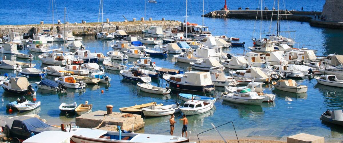 A marina with numerous small boats docked, a lighthouse in the background, and a rocky coastline.