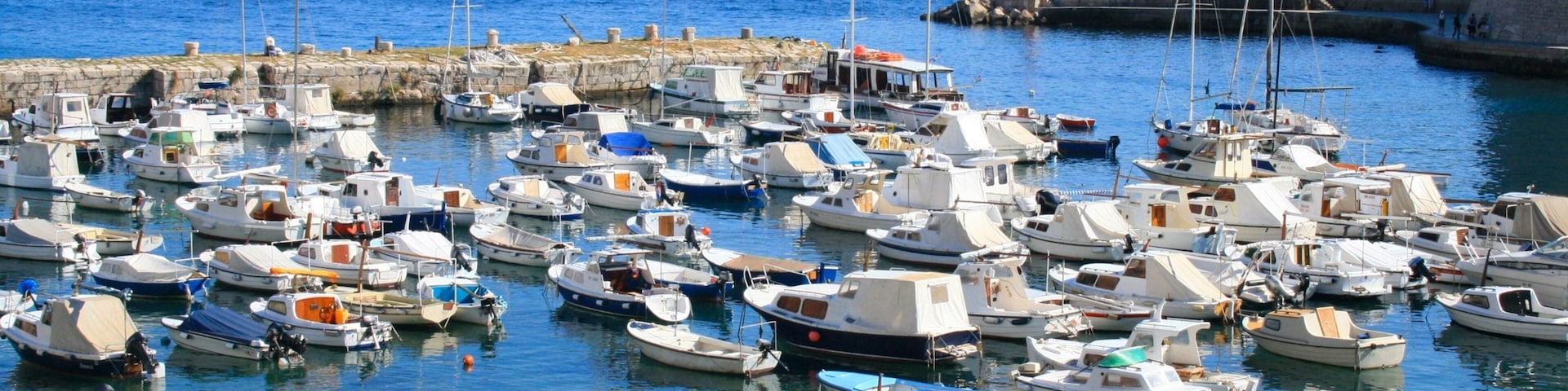 A marina with numerous small boats docked, a lighthouse in the background, and a rocky coastline.