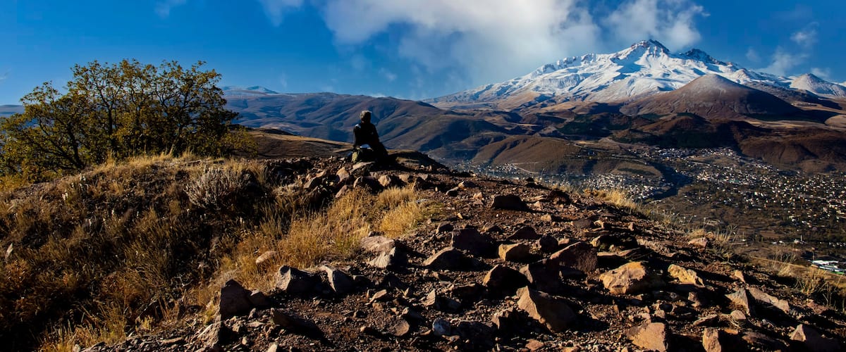 A person sitting on a rocky outcrop with a snow-capped mountain in the background.