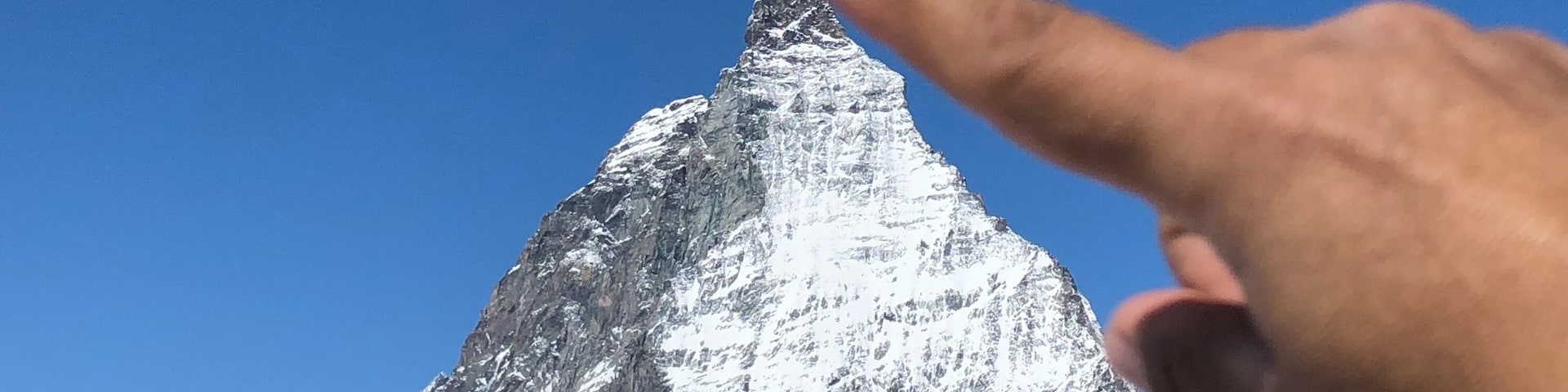 Eine Hand zeigt auf einen mit Schnee bedeckten Berggipfel unter blauem Himmel.
