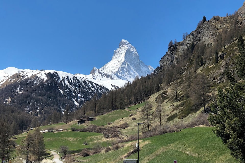 A snow-capped mountain with a clear blue sky, a green valley with trees, and a small building in the foreground.
