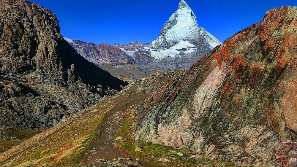 Un sentier de montagne qui mène à un sommet enneigé, avec un terrain accidenté de part et d’autre.