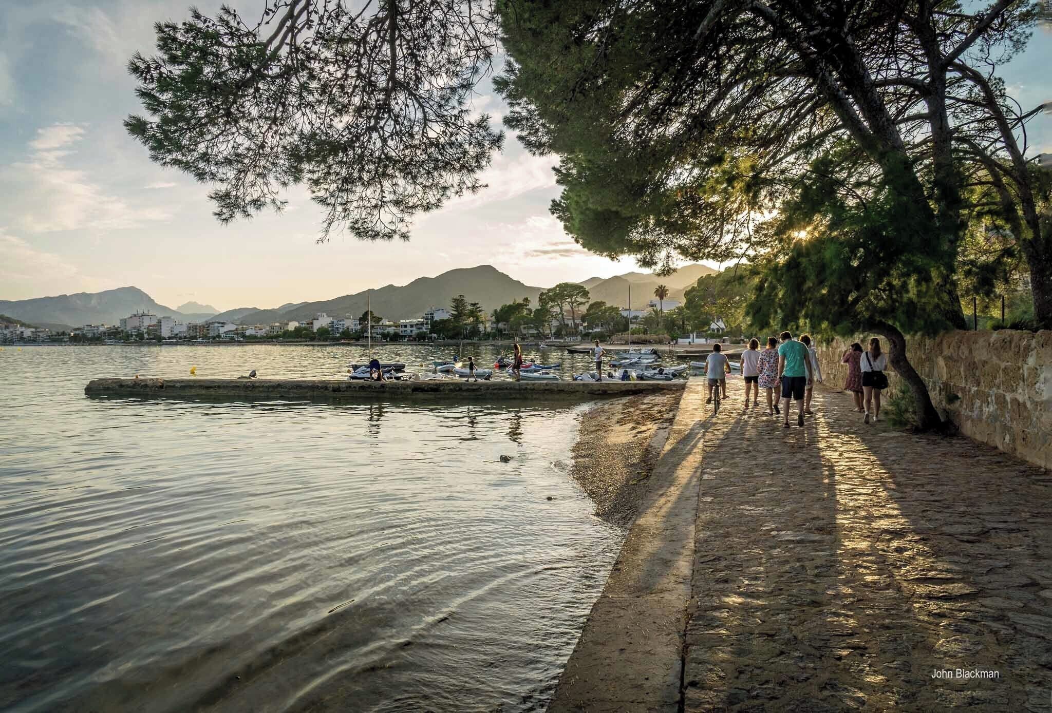 A waterfront promenade with people walking, boats docked, and mountains in the background.