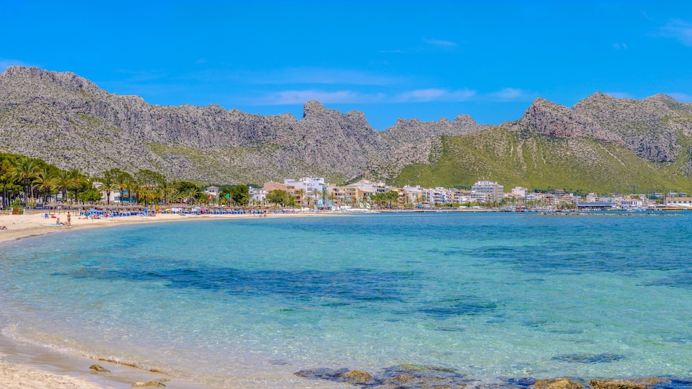 A beach with clear turquoise water, palm trees, and rocky mountains in the background.