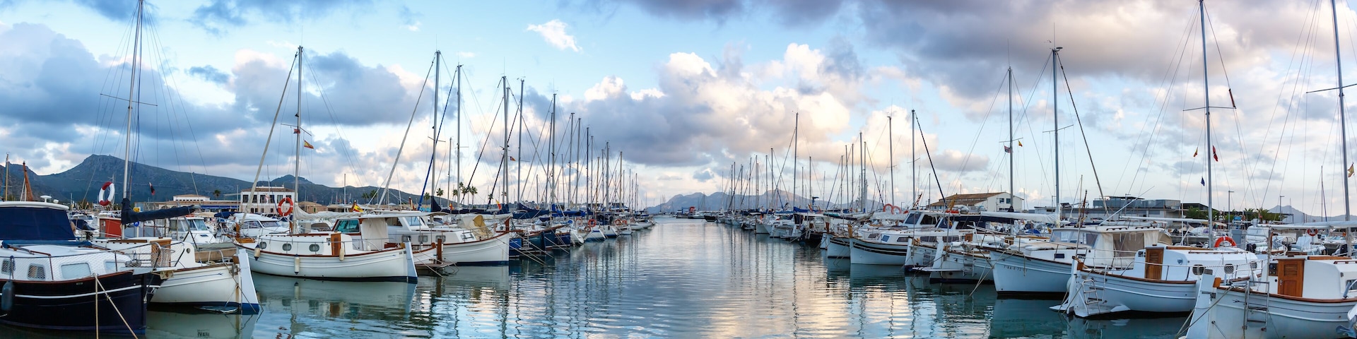 A marina with numerous sailboats docked in calm waters, surrounded by mountains and a partly cloudy sky.