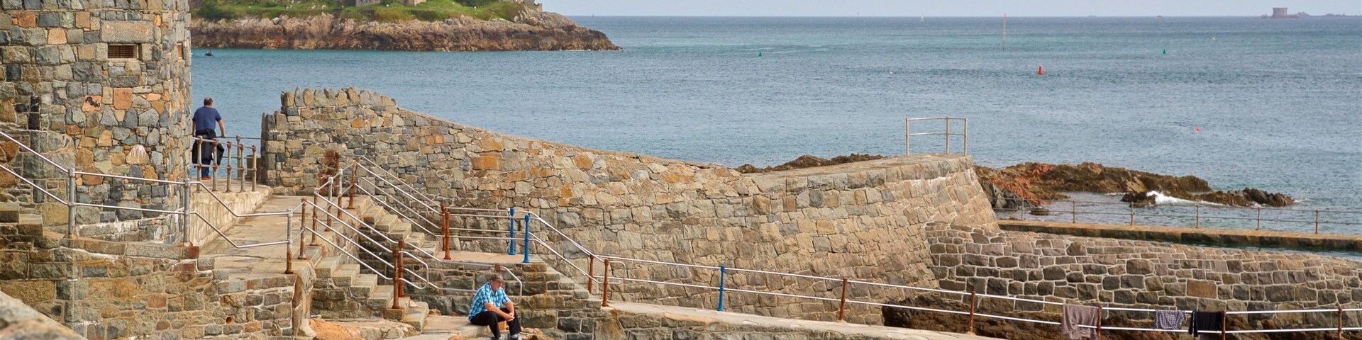 Uma fortificação costeira com paredes de pedra e uma escadaria que leva a uma plataforma com vista para o mar.