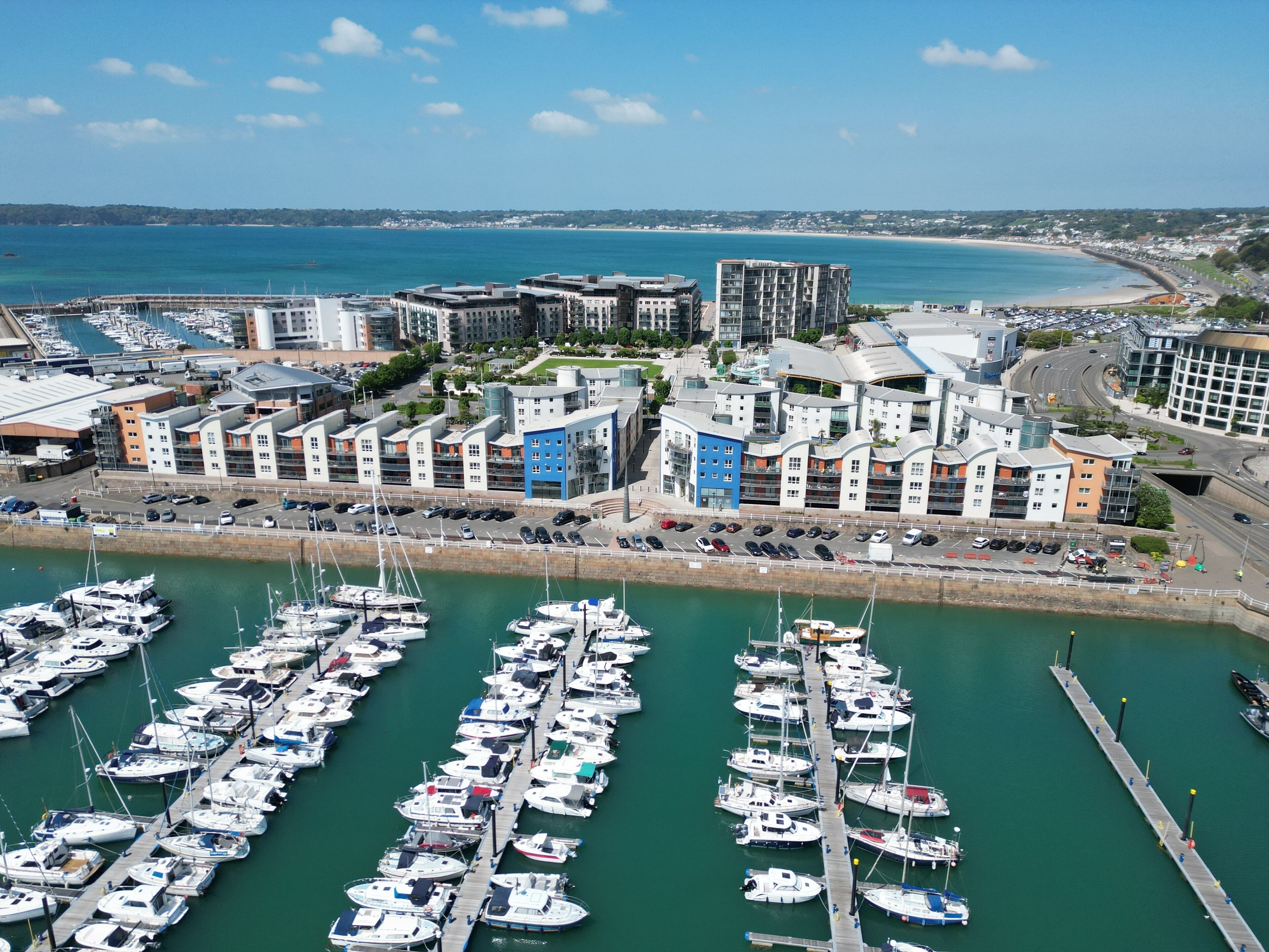 A marina with numerous boats docked, a coastal cityscape with modern buildings, and a clear blue sky.