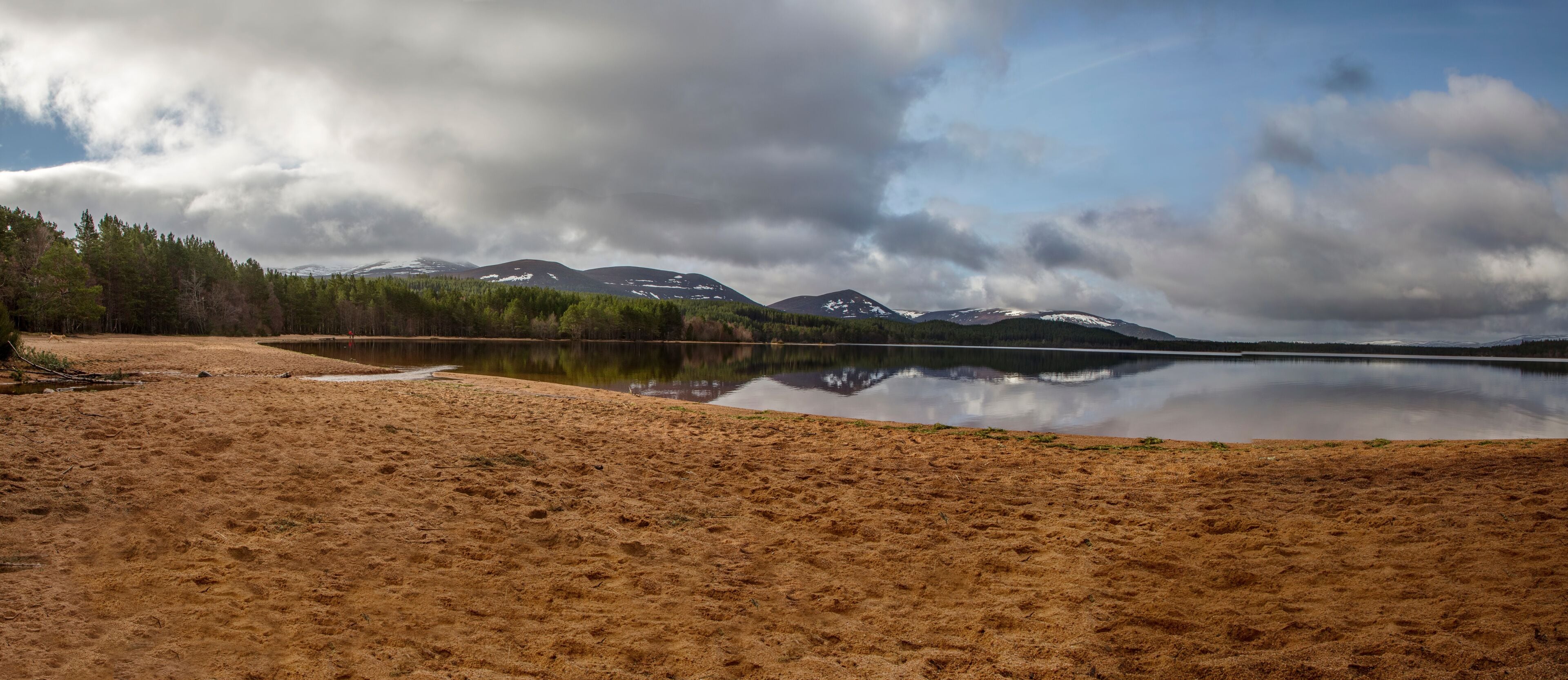Ein ruhiger See mit sandigem Ufer, umgeben von dichtem Wald und schneebedeckten Bergen unter bewölktem Himmel.