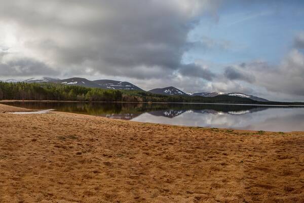 A calm lake with a sandy shore, surrounded by dense forest and snow-capped mountains under a cloudy sky.