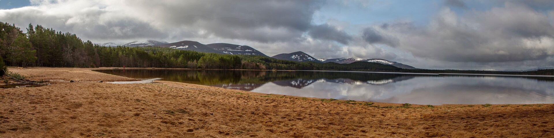 A calm lake with a sandy shore, surrounded by dense forest and snow-capped mountains under a cloudy sky.
