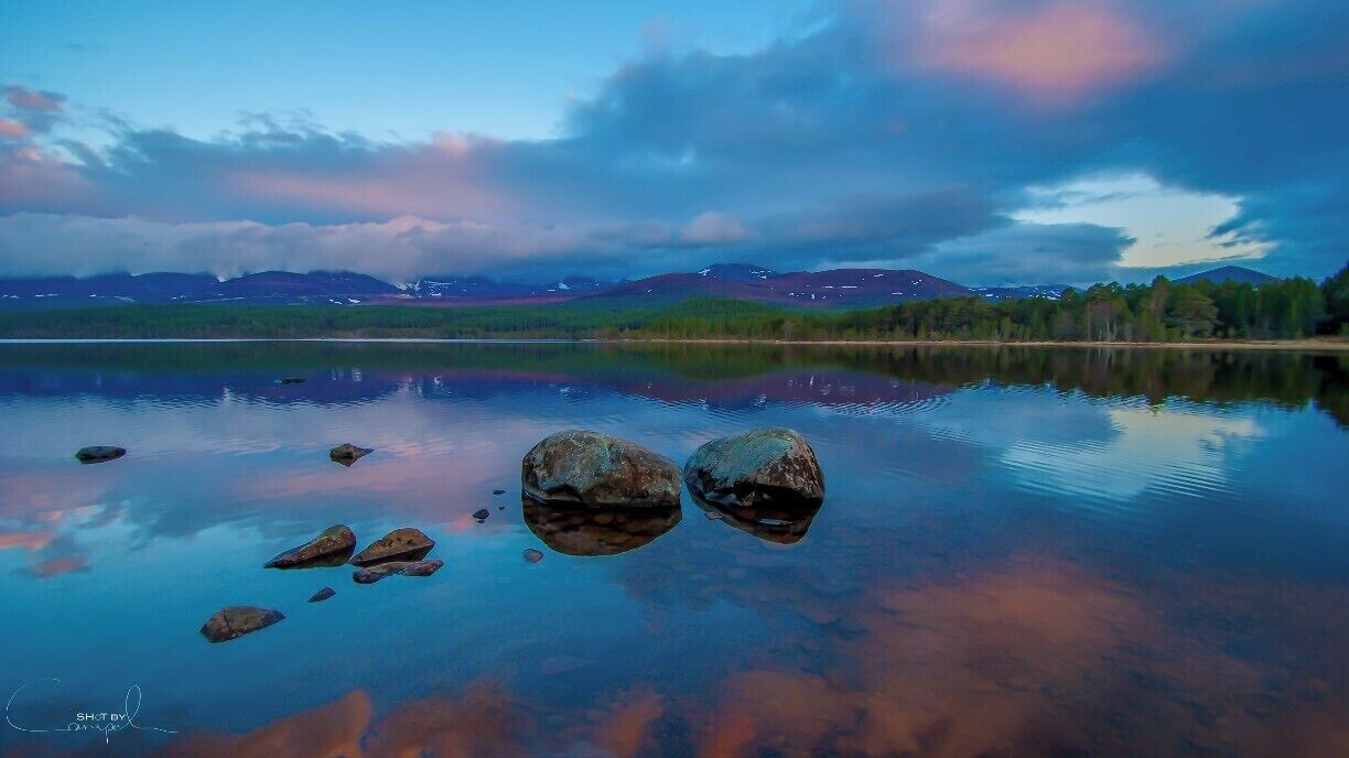 A calm lake with rocks in the foreground and mountains in the background.