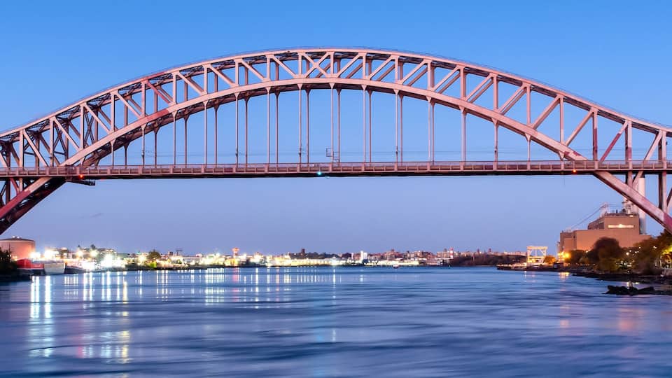A large steel bridge spanning a river with city lights in the background.
