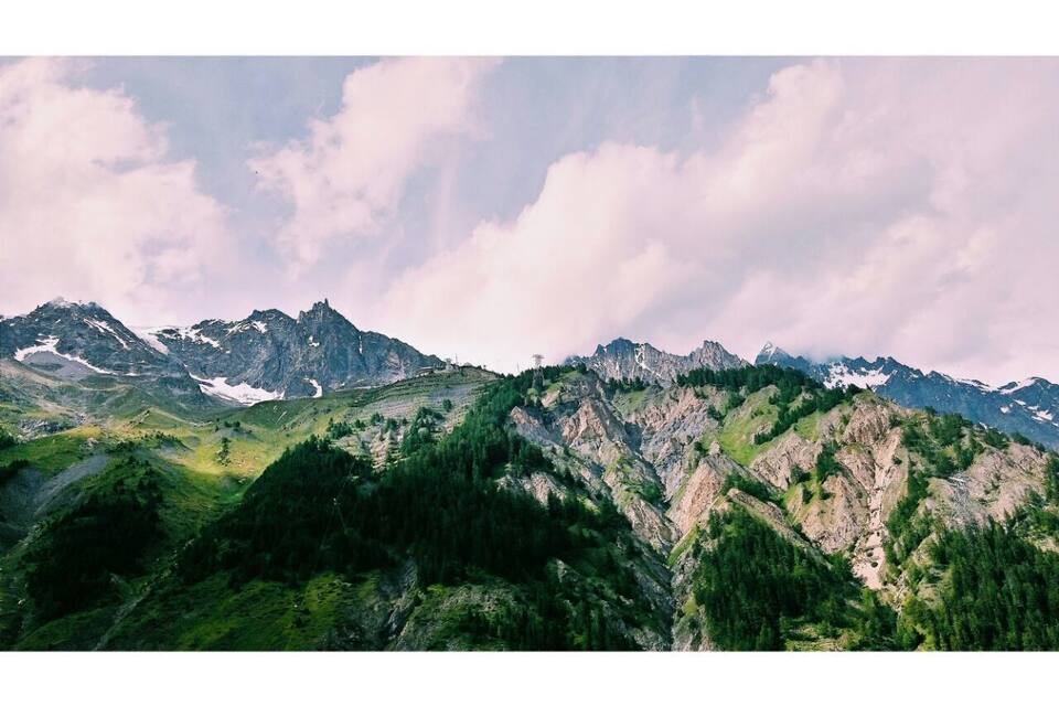 A mountain range with snow-capped peaks, dense forests, and a cloudy sky.