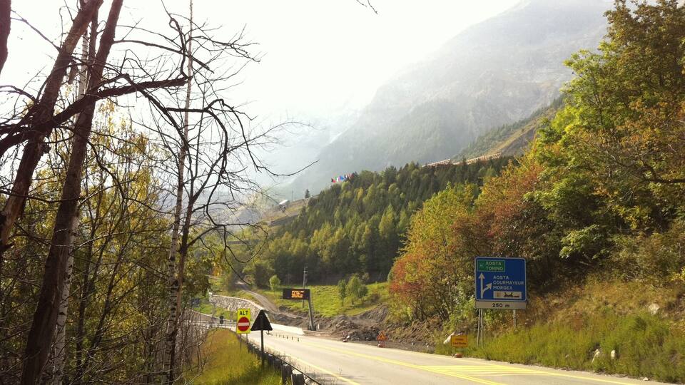 A mountain road with signs for Aosta and Courmayeur, surrounded by autumn foliage and a mountainous backdrop.