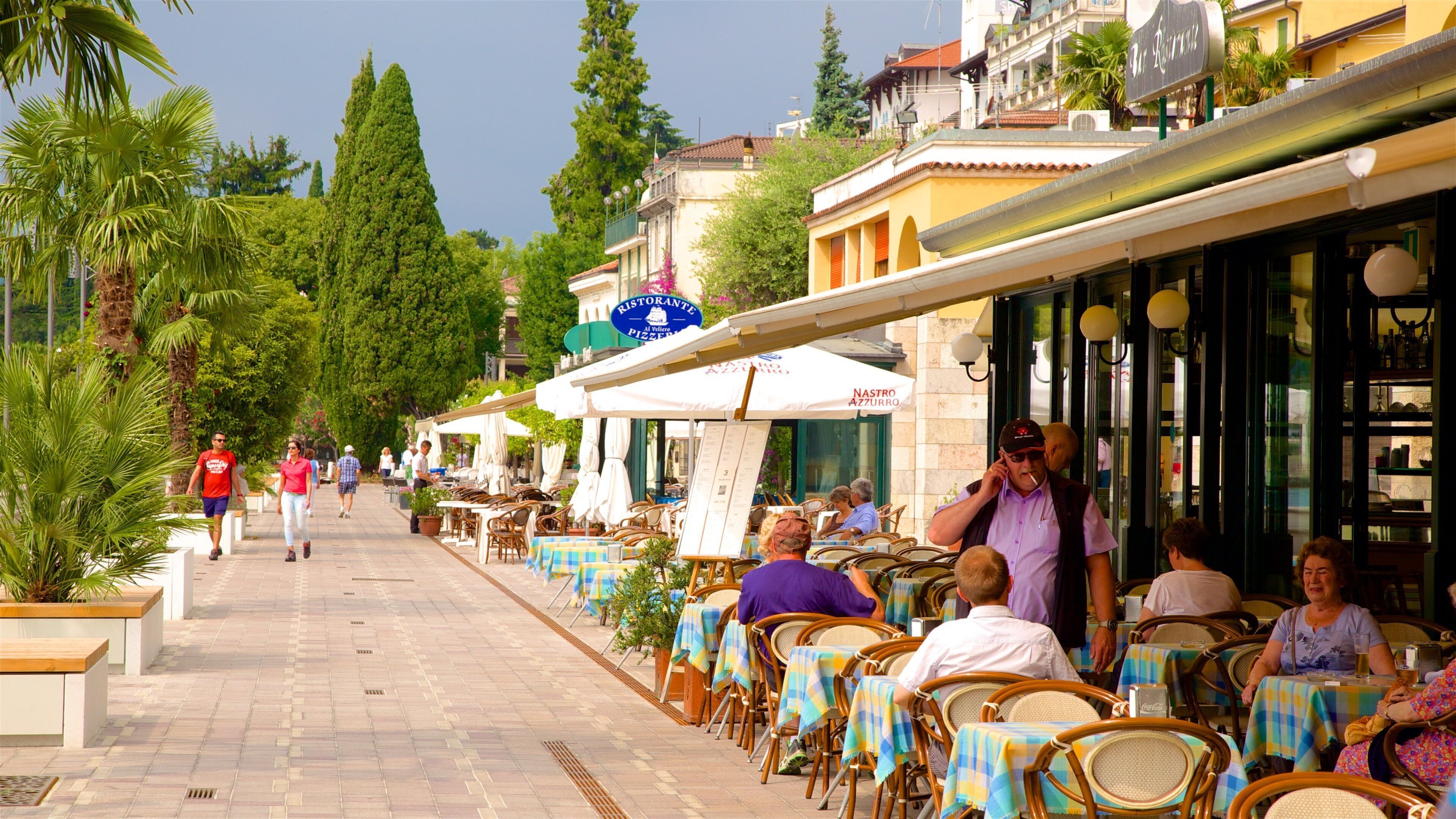 A sunny outdoor café with tables and chairs, people walking, and buildings in the background.