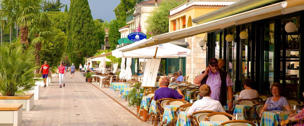 A sunny outdoor café with tables and chairs, people walking, and buildings in the background.