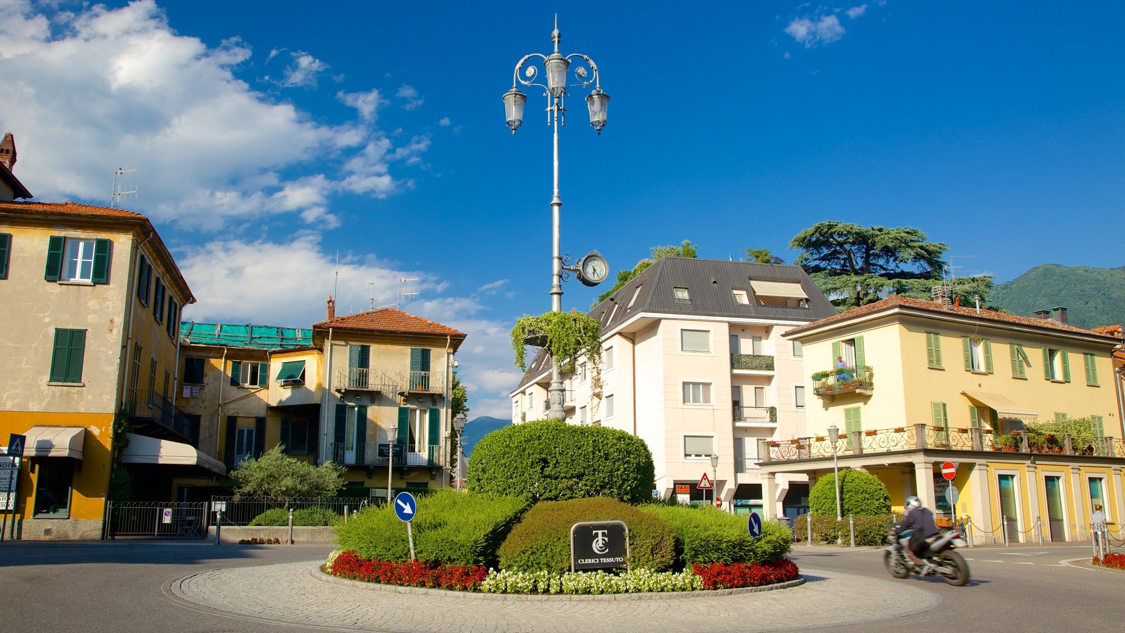 A roundabout with a clock tower, surrounded by buildings with green shutters, and a clear blue sky.