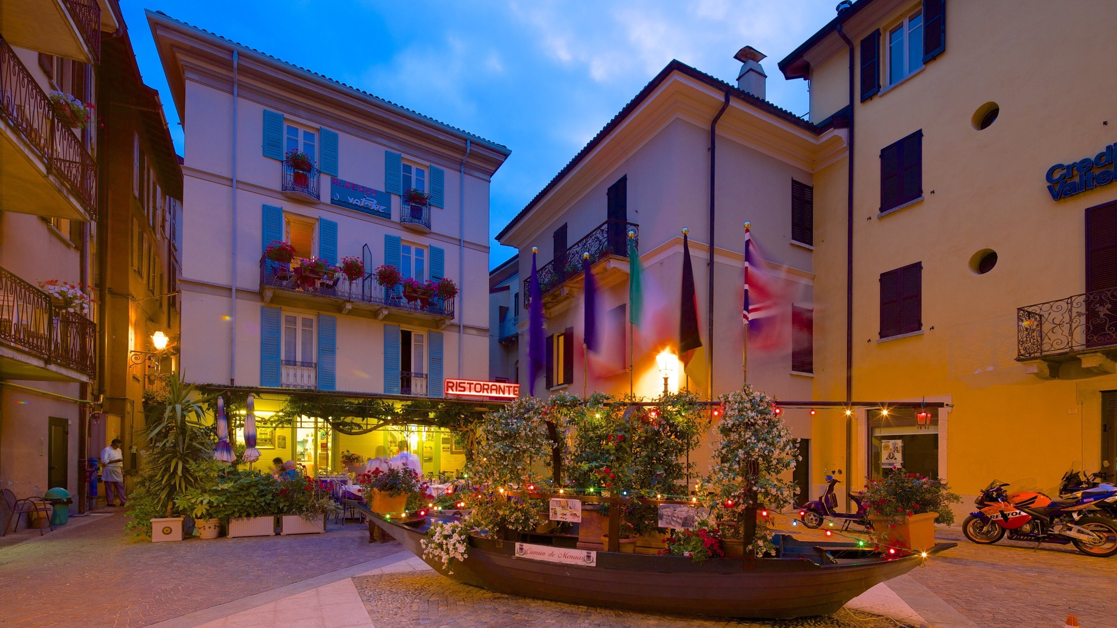 A plaza with a boat-shaped planter filled with flowers and lights, a restaurant named "Ristorante" with outdoor seating, and a building with balconies and flags.