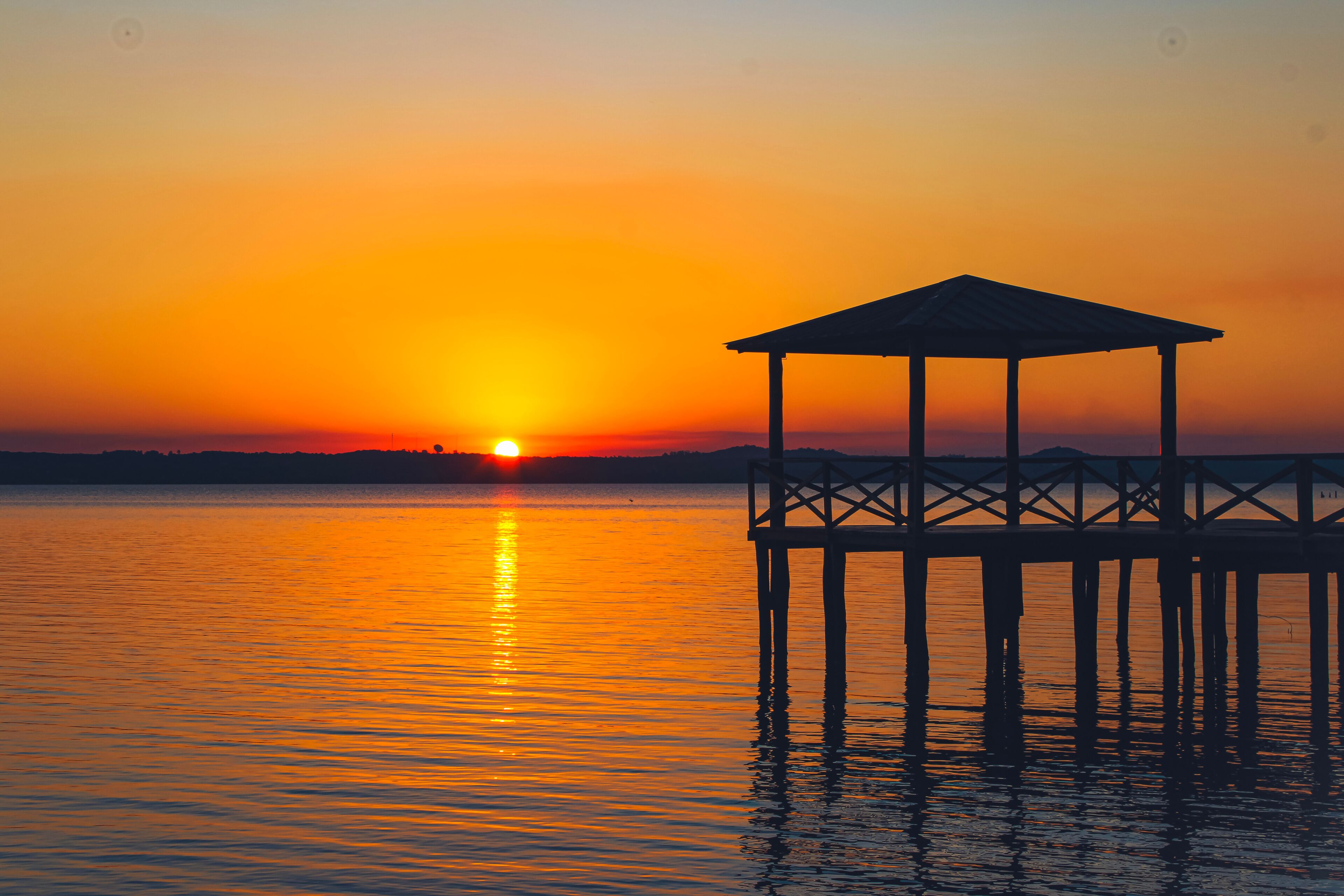 A sunset over a calm body of water with a wooden pier and a gazebo.
