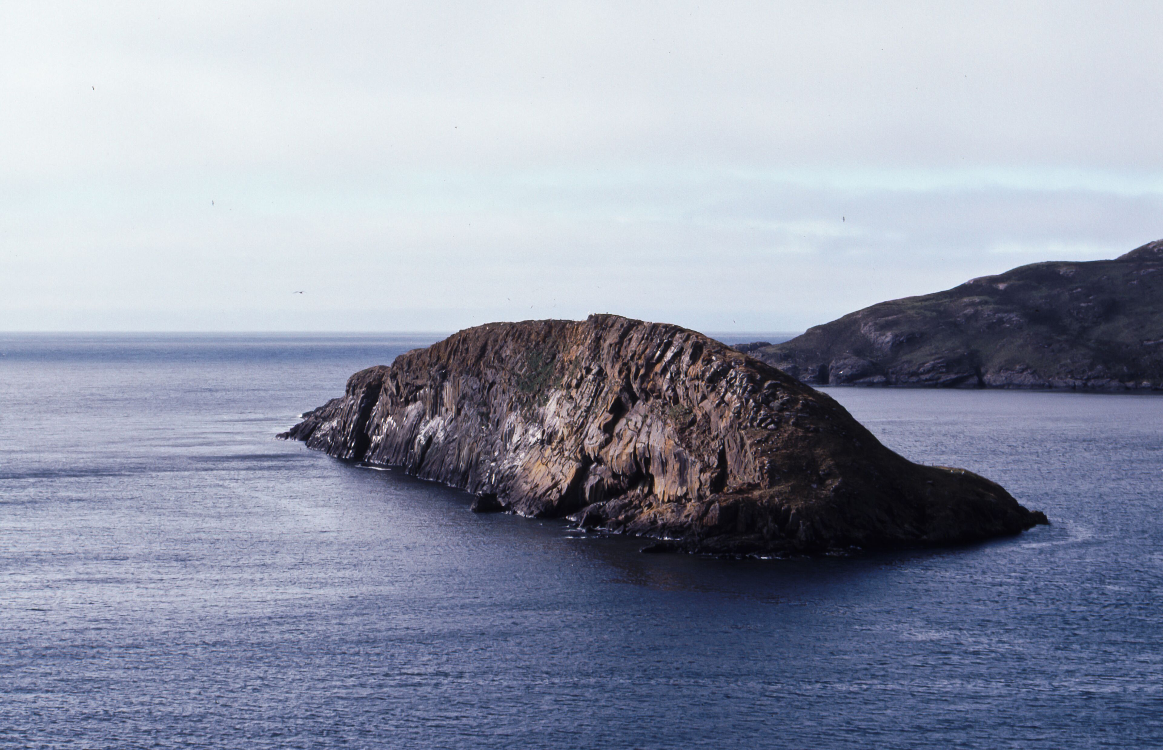 A large, rocky island in the ocean with a rugged coastline and a distant shoreline.
