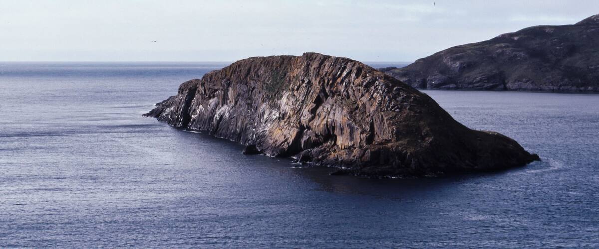 A large, rocky island in the ocean with a rugged coastline and a distant shoreline.