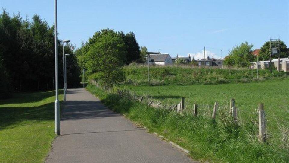 A paved pathway with street lamps, surrounded by grass and trees, leading towards a residential area.