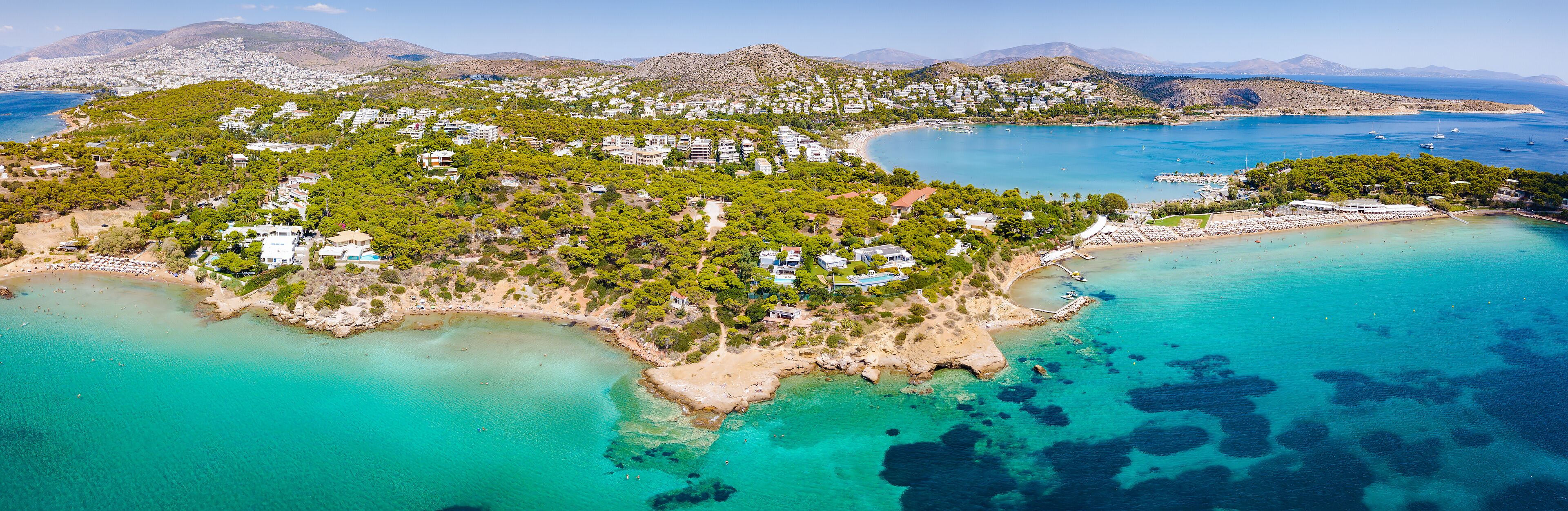 An aerial view of a coastal town with a beach, clear turquoise waters, and a backdrop of mountains.