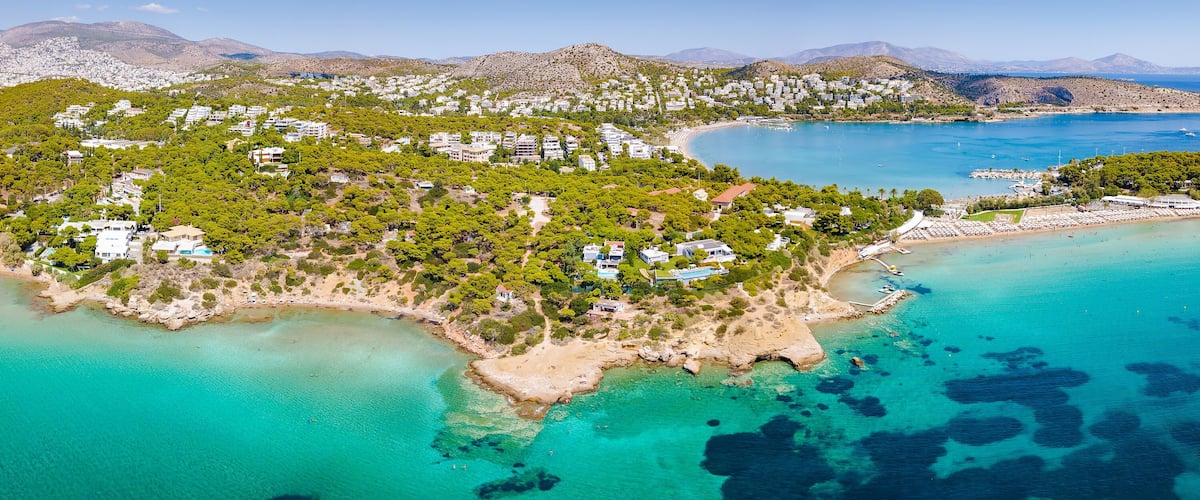 An aerial view of a coastal town with a beach, clear turquoise waters, and a backdrop of mountains.