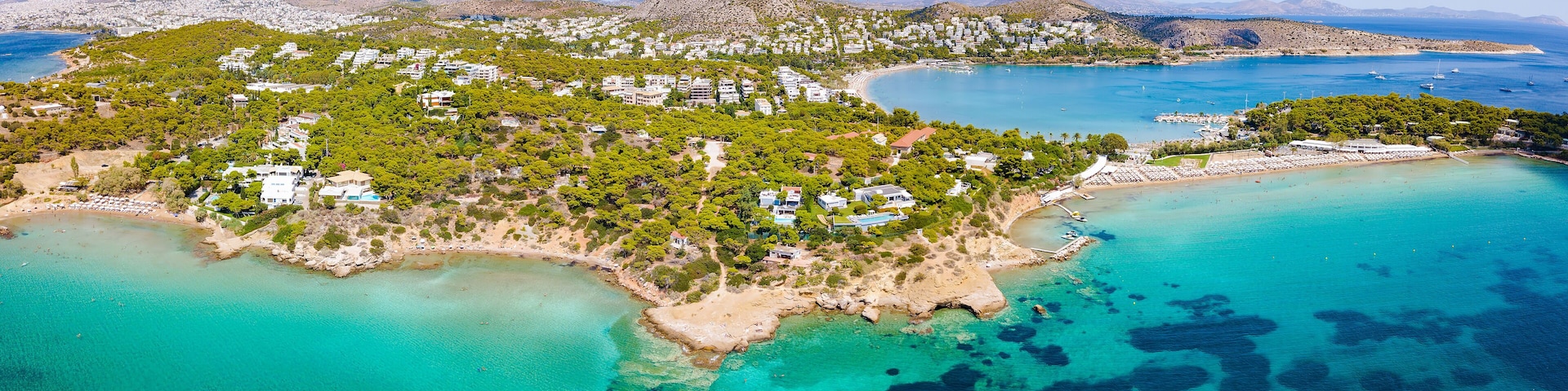 An aerial view of a coastal town with a beach, clear turquoise waters, and a backdrop of mountains.
