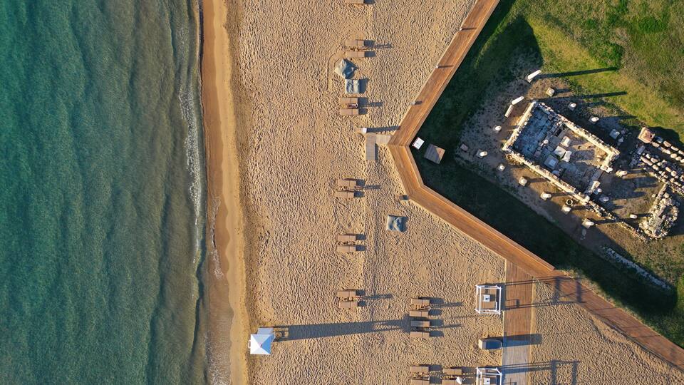 An aerial view of a beach with a wooden walkway and a fenced area with ruins.