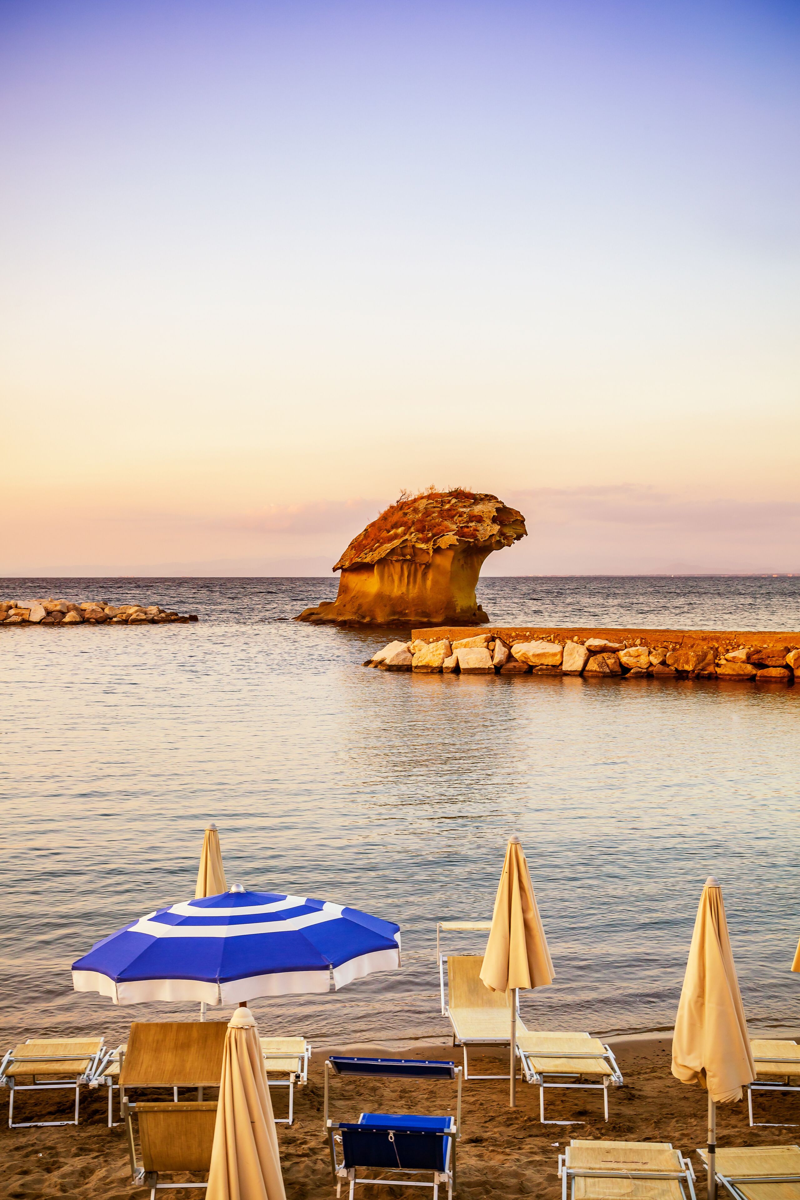 A beach with sun loungers, umbrellas, and a unique rock formation in the water.