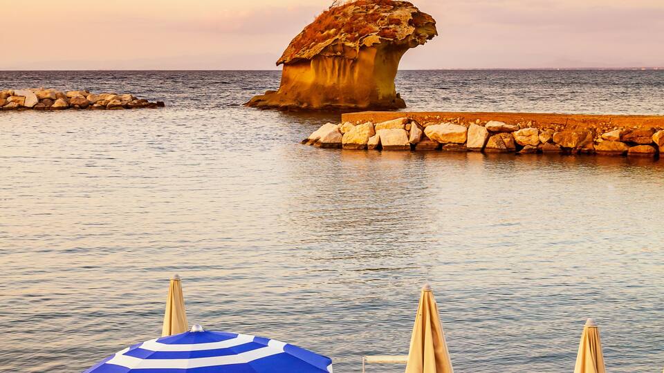 A beach with sun loungers, umbrellas, and a unique rock formation in the water.