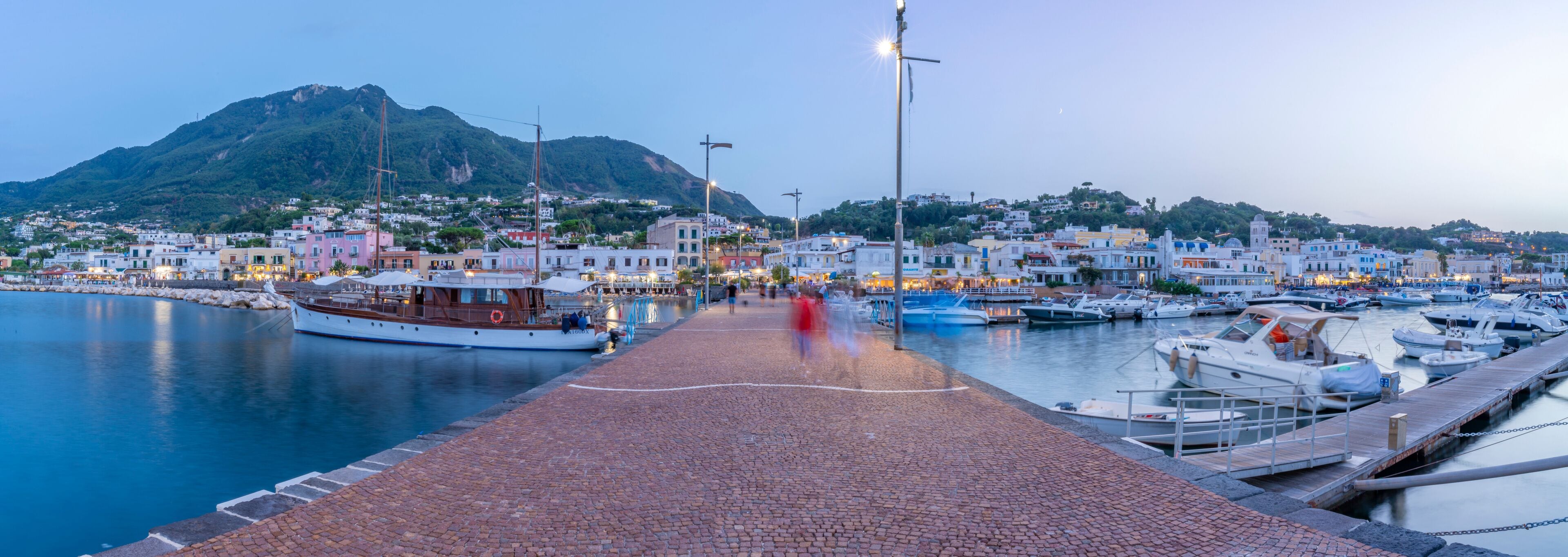 A harbor with boats docked, a cobblestone walkway, and buildings in the background.