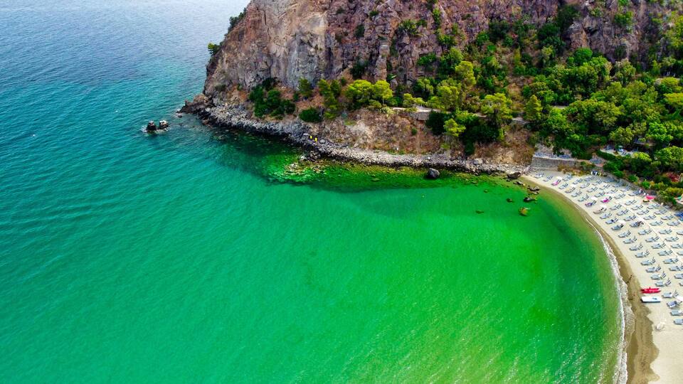 A coastal area with clear turquoise water, a rocky cliff, and a beach with sun loungers.
