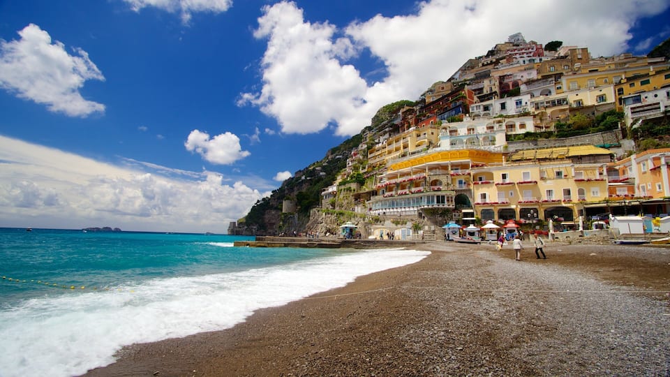 A pebble beach with colorful buildings on a hillside and the sea in the background.