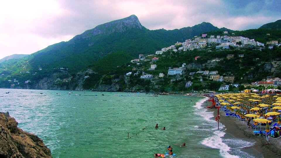 A coastal town with a beach, people swimming and sunbathing, and buildings on a hillside.