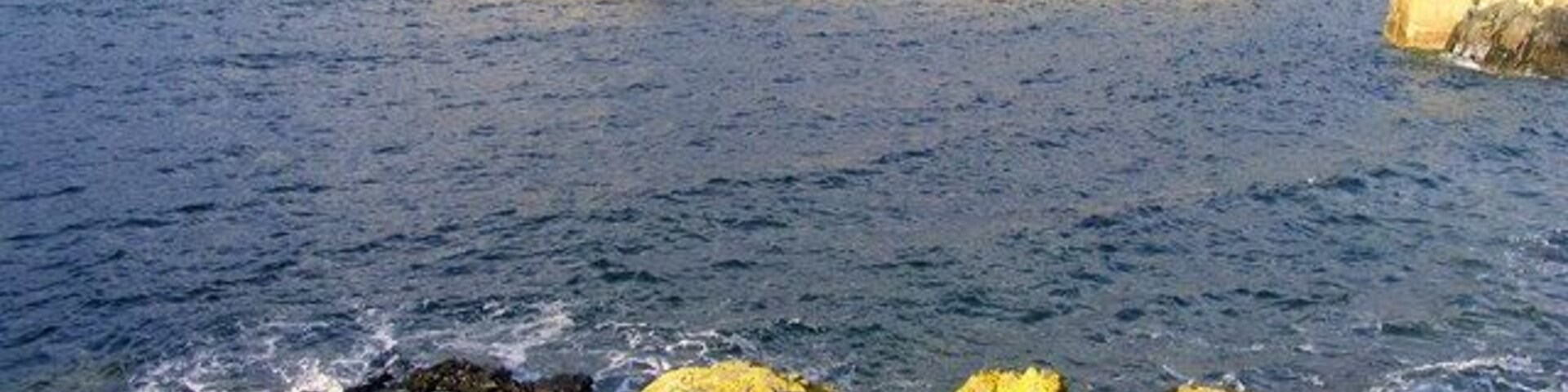 Un paisaje costero con un primer plano rocoso, una estructura de rompeolas y el mar.