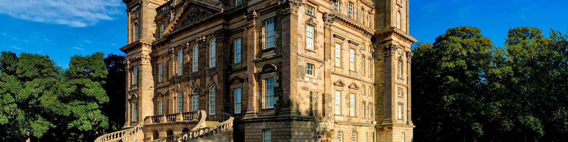 A large, historic building with a prominent tower, surrounded by lush green lawns and trees under a clear blue sky.