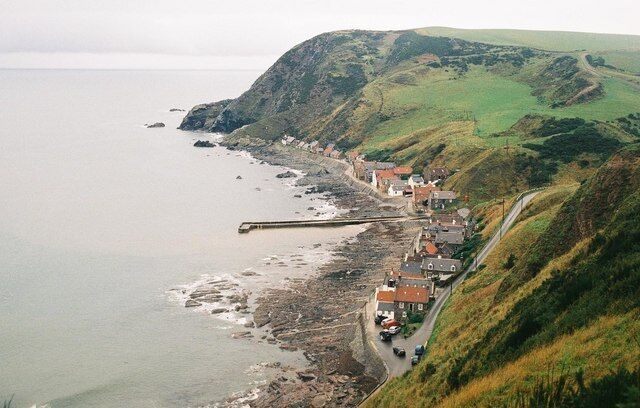 A coastal village with houses along the shoreline, a pier extending into the sea, and rolling hills in the background.