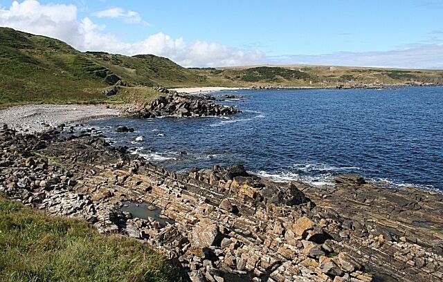 A rocky coastline with waves crashing onto the shore, grassy hills in the background, and a clear sky.