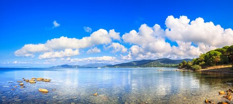 Un lac aux eaux limpides, parsemé de rochers, un voilier à l’horizon et un paysage montagneux sous un ciel bleu.