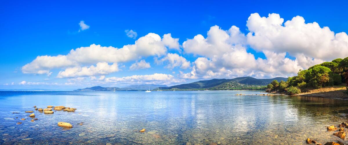 A clear lake with rocks, a distant sailboat, and a mountainous landscape under a blue sky.