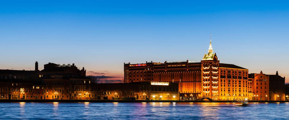 A grand illuminated building with a spire, a river in the foreground, and a city skyline in the background.