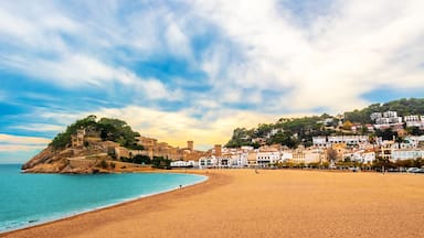 Un poble costaner amb una platja de sorra, una fortalesa històrica sobre un turó i edificis blancs al llarg de la costa.