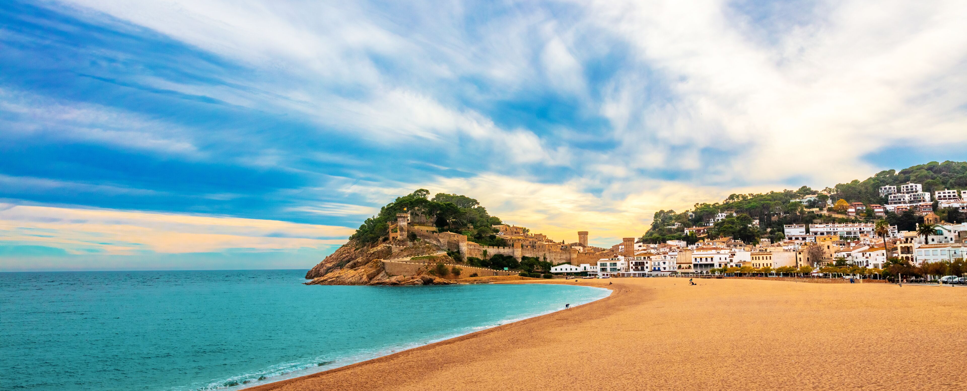 A coastal town with a sandy beach, a historic fort on a hill, and white buildings along the shore.