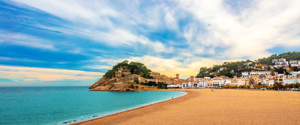 A coastal town with a sandy beach, a historic fort on a hill, and white buildings along the shore.